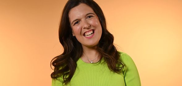 Rosie Jones smiles while wearing a neon green jumper and standing against a beige background.