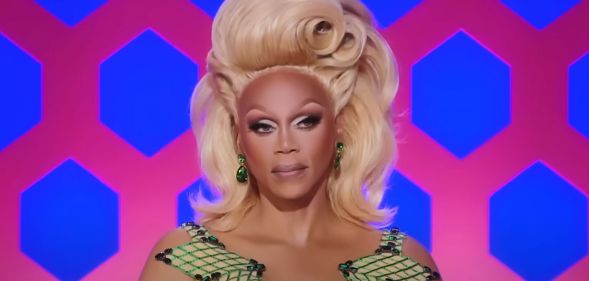 A still of RuPaul from RuPaul's Drag Race wearing a green patterned dress and blonde wig.
