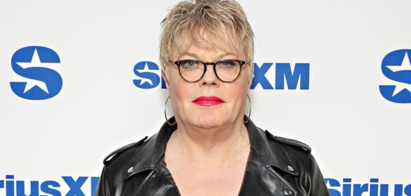 Suzy Izzard wears black rimmed glasses, a black leather jacket, and hooped earrings as she poses on a red carpet.