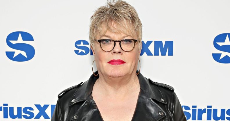 Suzy Izzard wears black rimmed glasses, a black leather jacket, and hooped earrings as she poses on a red carpet.