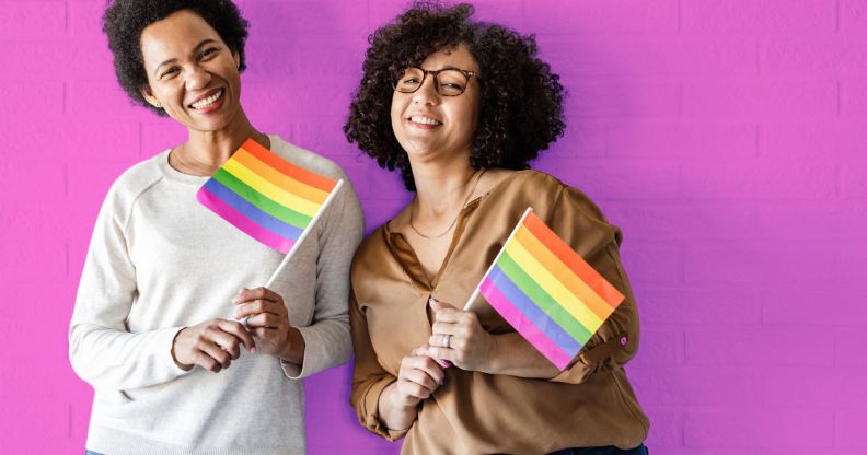 This is an image of two queer women holding the Pride flag on a pink wall.