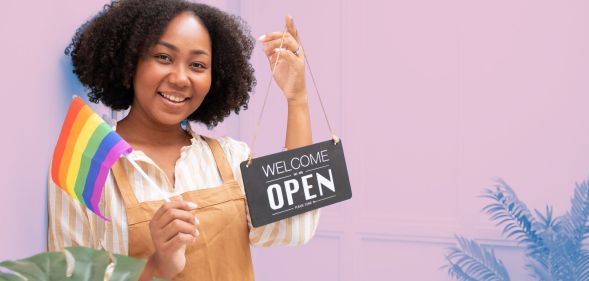 This is an image of a Black woman holding an "we're open" sign in one hand and a Pride flag in the owner. She is wearing a brown apron.