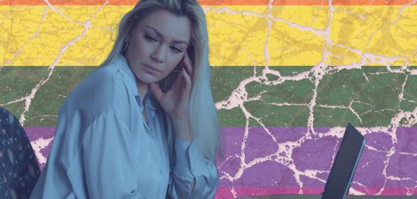 this is a stylized image of a woman looking unhappy or distressed at a computer. She is in a blue tint and behind her is the pride flag, but it is cracked.
