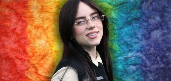 Billie Eilish looks off to the right and smiles. She is wearing glasses and a beige shirt and black vest. She is against a rainbow background.
