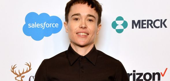 Elliot Page smiles on a red carpet while wearing a dark buttoned shirt.