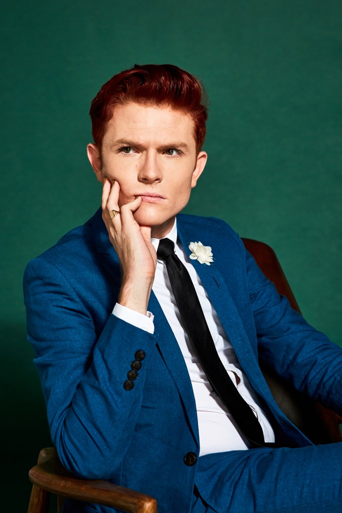 Rhys Nicholson: Huge Big Party Congratulations! Ryan is in a blue suit sat on a brown chair against a green background. He's resting his face on his hand, looking seriously.