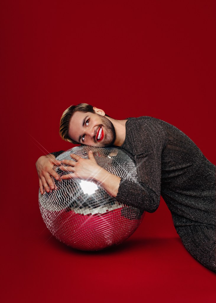 Will Owen in a sparkly look with red lipstick hugging a mirrorball.