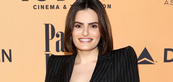 Nava Mau poses and smiles on a red carpet wearing a black pin-striped suit.