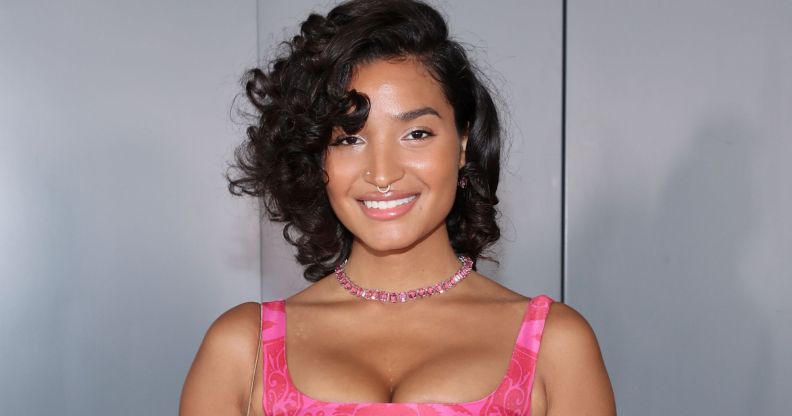 Indya Moore smiles and poses while wearing a pink dress and standing against a grey background.