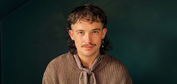 Singer songwriter Freddie Lewis poses against a green wall while wearing a brown jumper