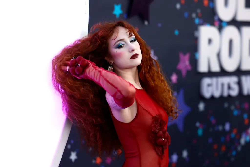 Chappell Roan attends the premiere of Netflix's Olivia Rodrigo: GUTS World Tour. She's brushing her long red curly hair off her shoulder, fearing a sheer red dress.