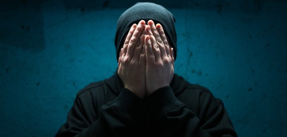 Color image depicting an adult man wearing a black beanie hat in an underground subway tunnel. His expression is one of despair, sadness and depression. His hands are covering his face in a gesture of bleak desperation and hopelessness.