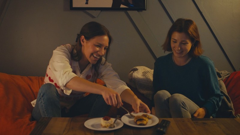 The Holiday Club still: two women sharing desert while sat together on a sofa