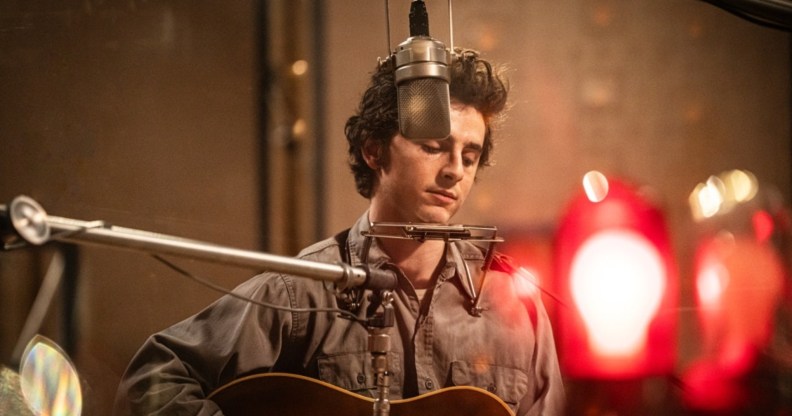 A Complete Unknown still: Timothée Chalamet as Bob Dylan in a studio recording. He's got a harmonica stand around his neck and playing the guitar.