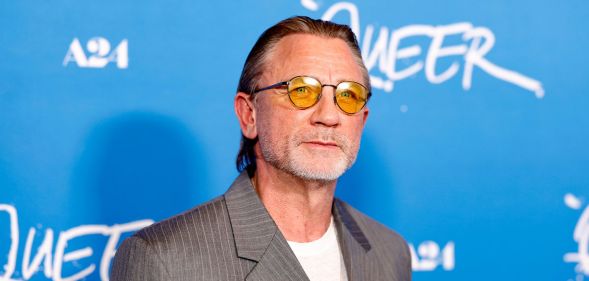 Daniel Craig in front of a blue background wearing orange lens glasses, a grey suit and white t-shirt