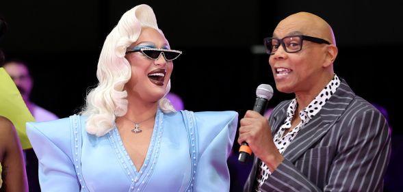 RuPaul holds a microphone to her mouth while The Vivienne looks at her smiling.