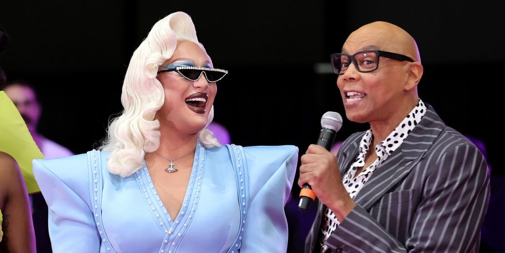 RuPaul holds a microphone to her mouth while The Vivienne looks at her smiling.