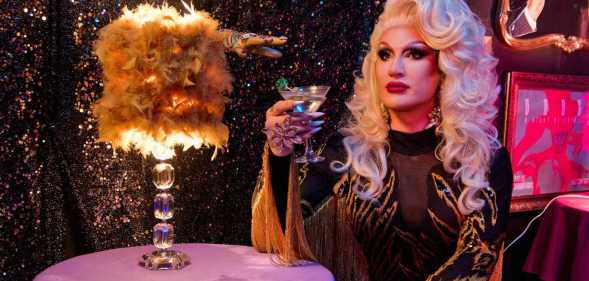 The Vivienne in a blonde wig while holding a cocktail glass.