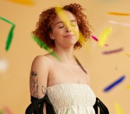 Smiling young redhead non-binary person standing amidst falling confetti against beige background