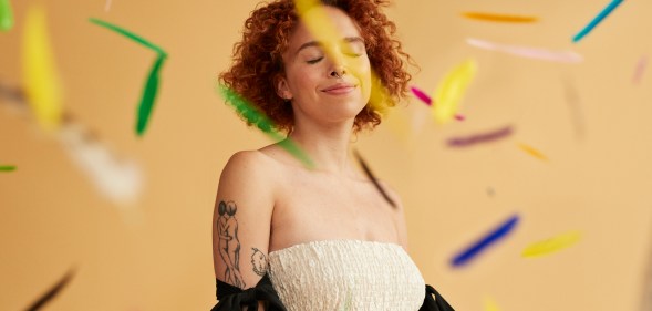 Smiling young redhead non-binary person standing amidst falling confetti against beige background