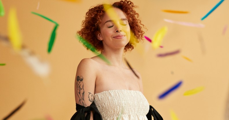 Smiling young redhead non-binary person standing amidst falling confetti against beige background