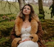 Charlotte Church looking pensive while perching in a forest in a white dress and brown fluffy coat.