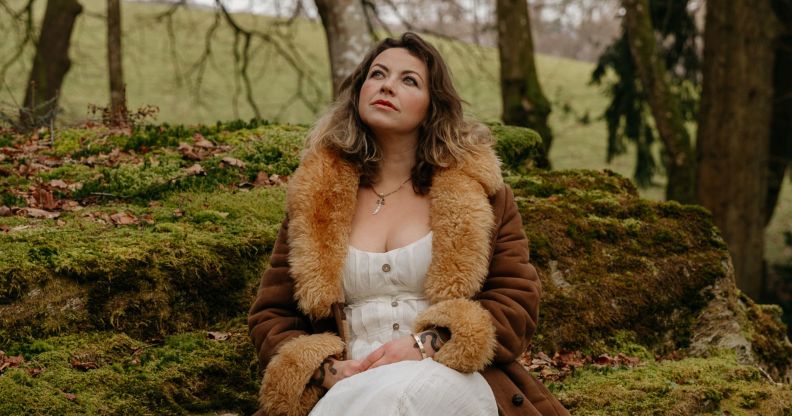 Charlotte Church looking pensive while perching in a forest in a white dress and brown fluffy coat.
