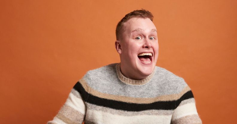 Comedian Josh Jones beams with his mouth open. He is in a grey black and beige striped jumper and sitting against an orange background.
