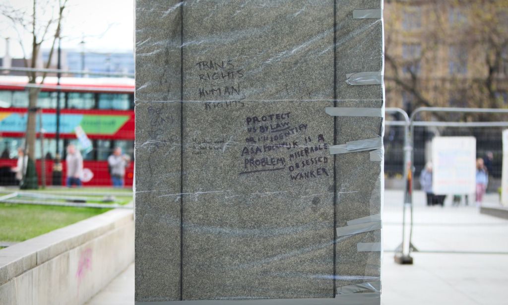 Protective film wrapped over the base of a statue where trans rights slogans have been tagged.