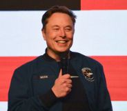 Elon Musk stands with a microphone in front of an American flag background