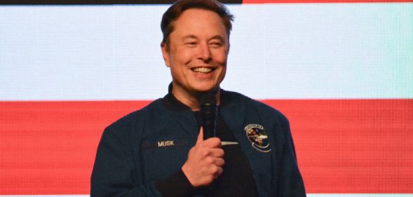 Elon Musk stands with a microphone in front of an American flag background