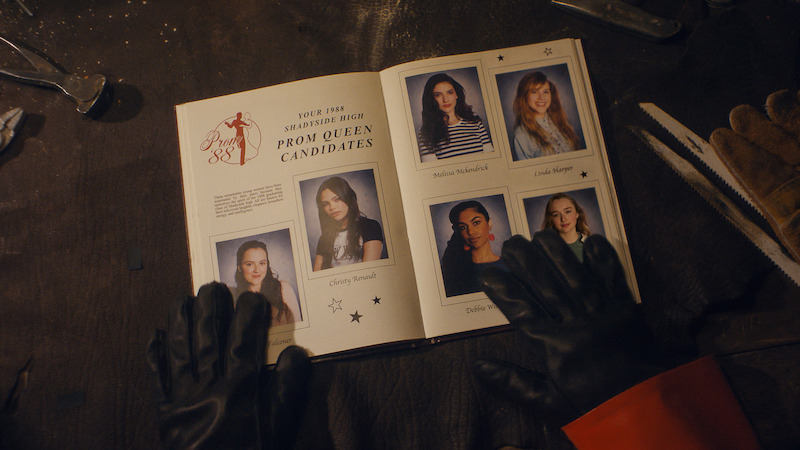Fear Street: Prom Queen still: a prom book of the 5 prom queen candidates. A pair of gloved hands is holding the book open with a saw on the table