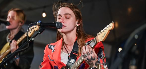 Julien Baker singing into a mic with her eyes closed and her brown hair billowing in the wind above her head. She is holding a guitar and wearing a red blazer and white top.