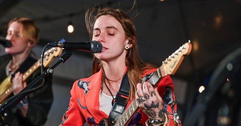 Julien Baker singing into a mic with her eyes closed and her brown hair billowing in the wind above her head. She is holding a guitar and wearing a red blazer and white top.