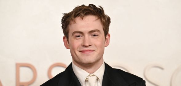 Kit Connor smiles at a red carpet event while wearing a cream t-shirt, black blazer, against a cream background.
