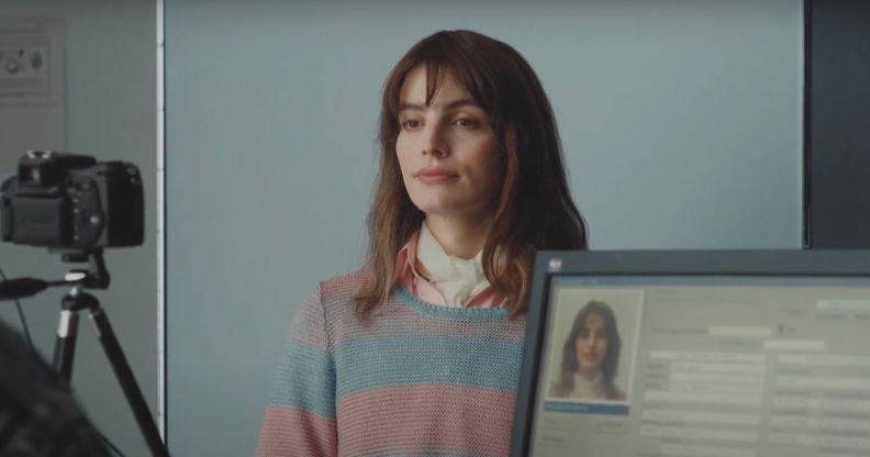 Lux Pascal in a blue and pink striped jumped in a scene in new film Miss Carbon. She is stood in front of a blue wall and behind a computer screen and camera.