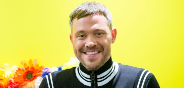 Will Young in a black and white jacket smiling while posing against a yellow and orange background.