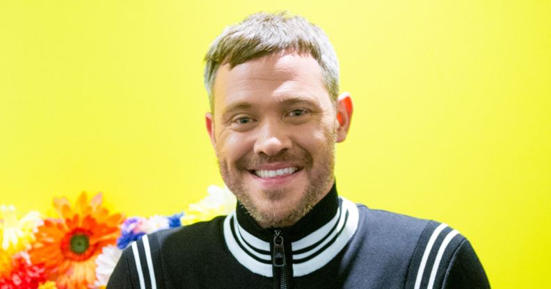 Will Young in a black and white jacket smiling while posing against a yellow and orange background.