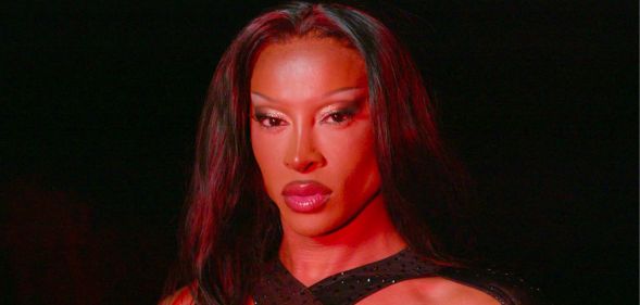 Drag star Tayce in full drag standing in a dark room with a red light shining on her face.