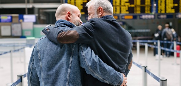 A gay couple walking and embracing each other closely while making their way through an airport, on their way to the check-in before catching a flight together.