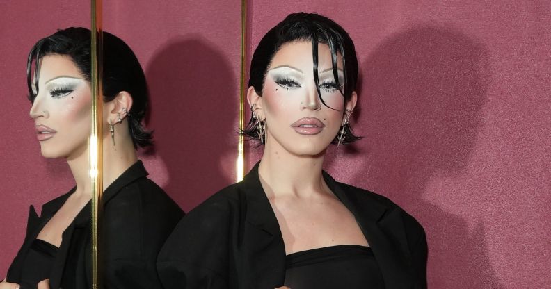 Drag Race winner Aquaria stands against a mirror while wearing a black outfit and sicked back black hair