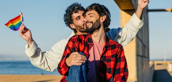 Mid-adult male gay couple embracing while holding LGBTQ+ flags outdoors in Barcelona in Spain.