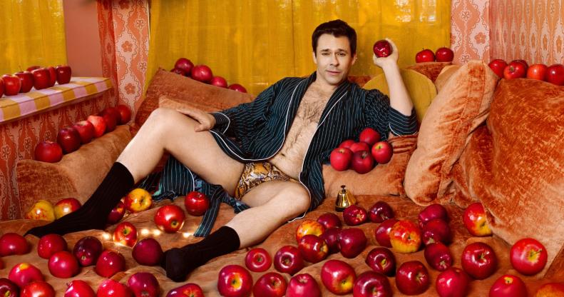Bryan Safi wearing leopard print underpants and an open robe. He is laying on a sofa covered in apples.