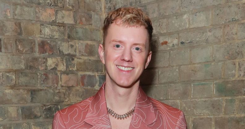 James Bar in a peach blazer and necklace smiling while posing against a brick wall.