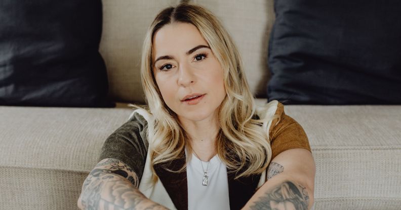 Lucy Spraggan poses while sitting in a green and brown shirt, white t-shirt, and blonde hair. She is sat on the floor in front of a sofa.