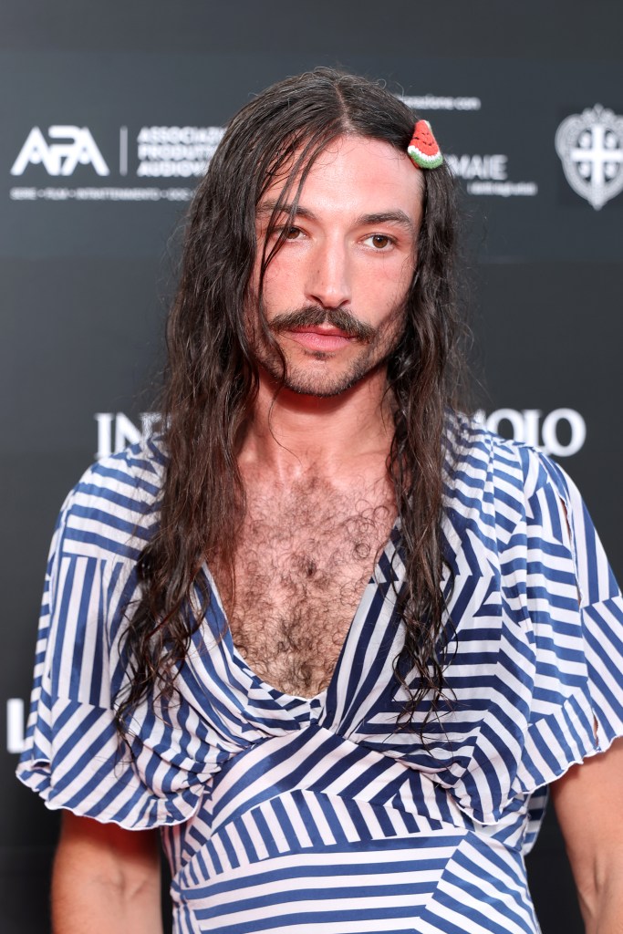 Ezra Miller attends the Filming Italy. They are wearing a blue and white stripey dress. A watermelon clip in their long brown wavy hair.