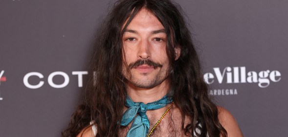 Ezra Miller attends Filming Italy 2025 They have long wavy brown hair over their shoulders. A silk blue necktie, gold beaded necklace and white vest.