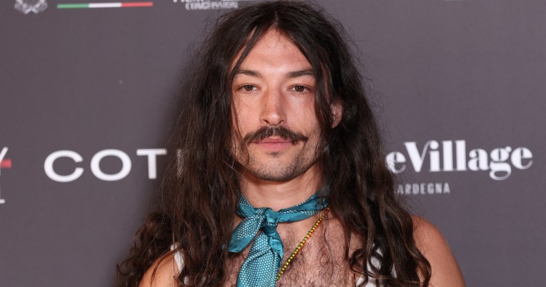 Ezra Miller attends Filming Italy 2025 They have long wavy brown hair over their shoulders. A silk blue necktie, gold beaded necklace and white vest.