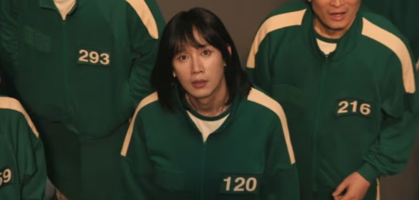 Squid Game character Player 120 looking up towards the camera, surrounded by other contestants.