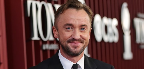 Tom Felton attends The 78th Annual Tony Awards at Radio City Music Hall. He's wearing a white shirt and black suit against a red Tony Awards background.